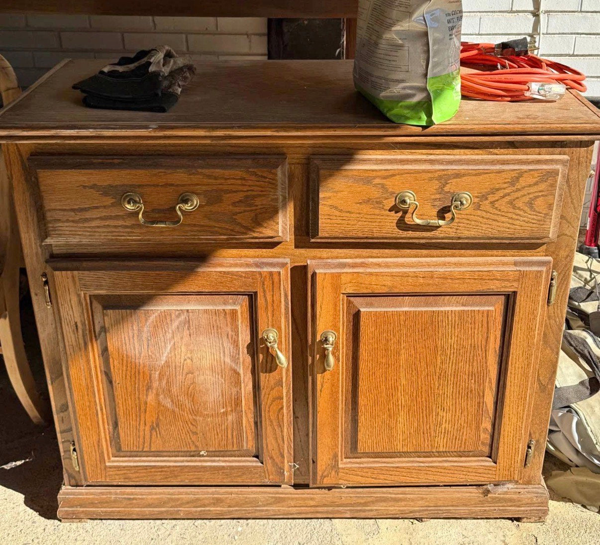 Old, worn wooden cabinet with drawers and doors. Areas are damaged and sanded, hardware is askew & piece appears to be sitting in a discard area.